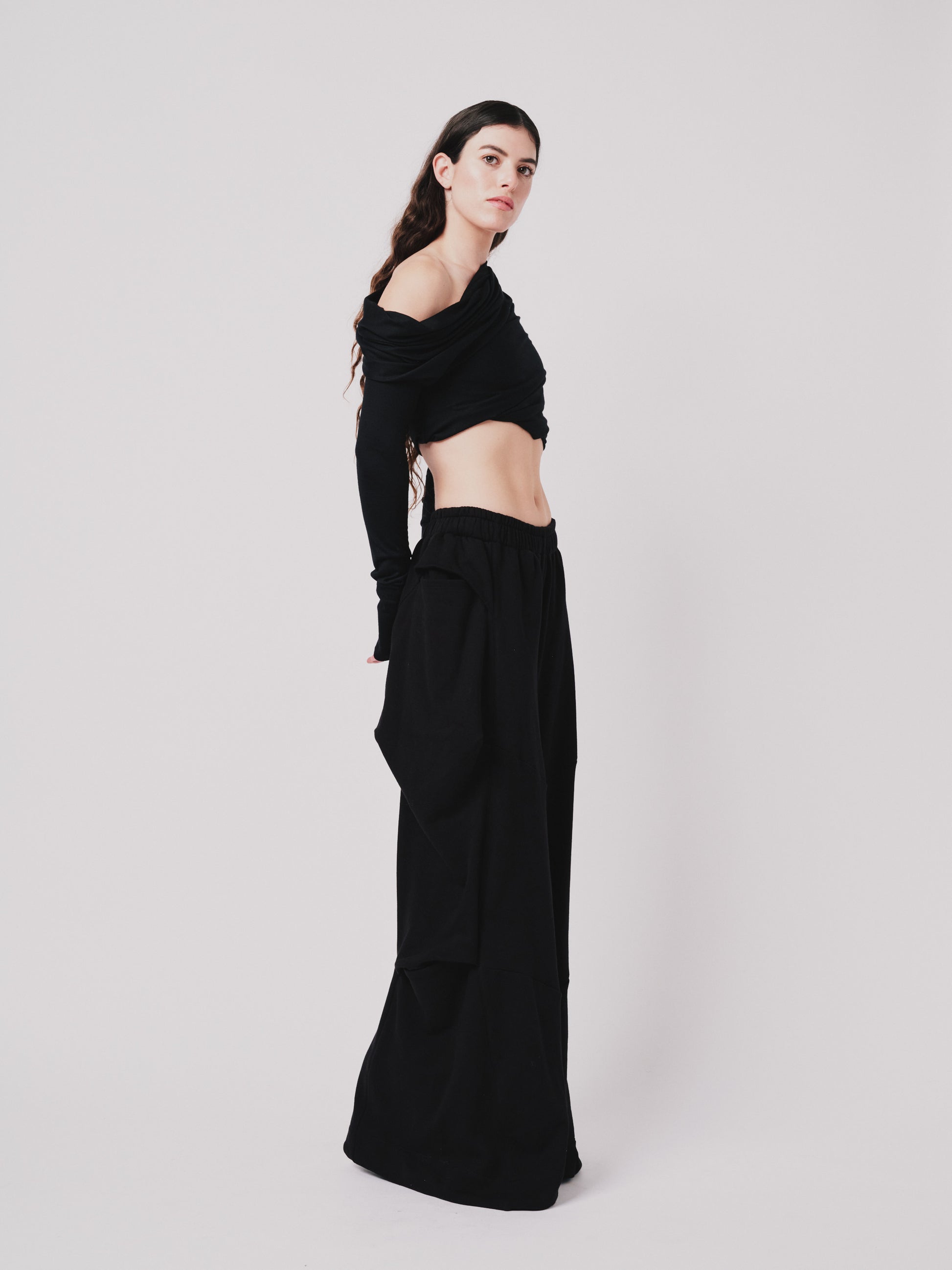 Woman wearing off the shoulder draped top. Cropped in black merino wool on white background. Side view