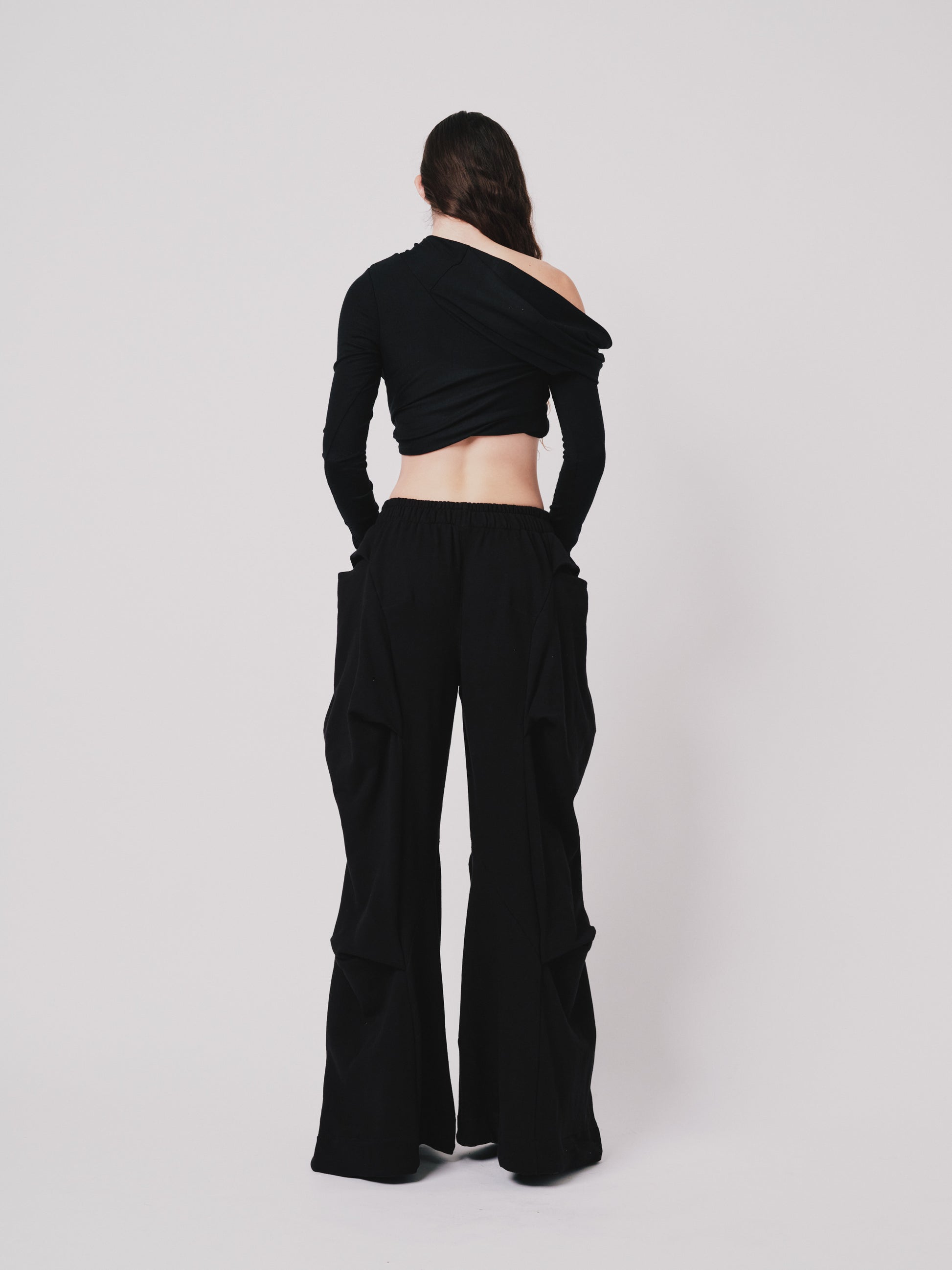 Woman wearing off the shoulder draped top. Cropped in black merino wool on white background. Back view
