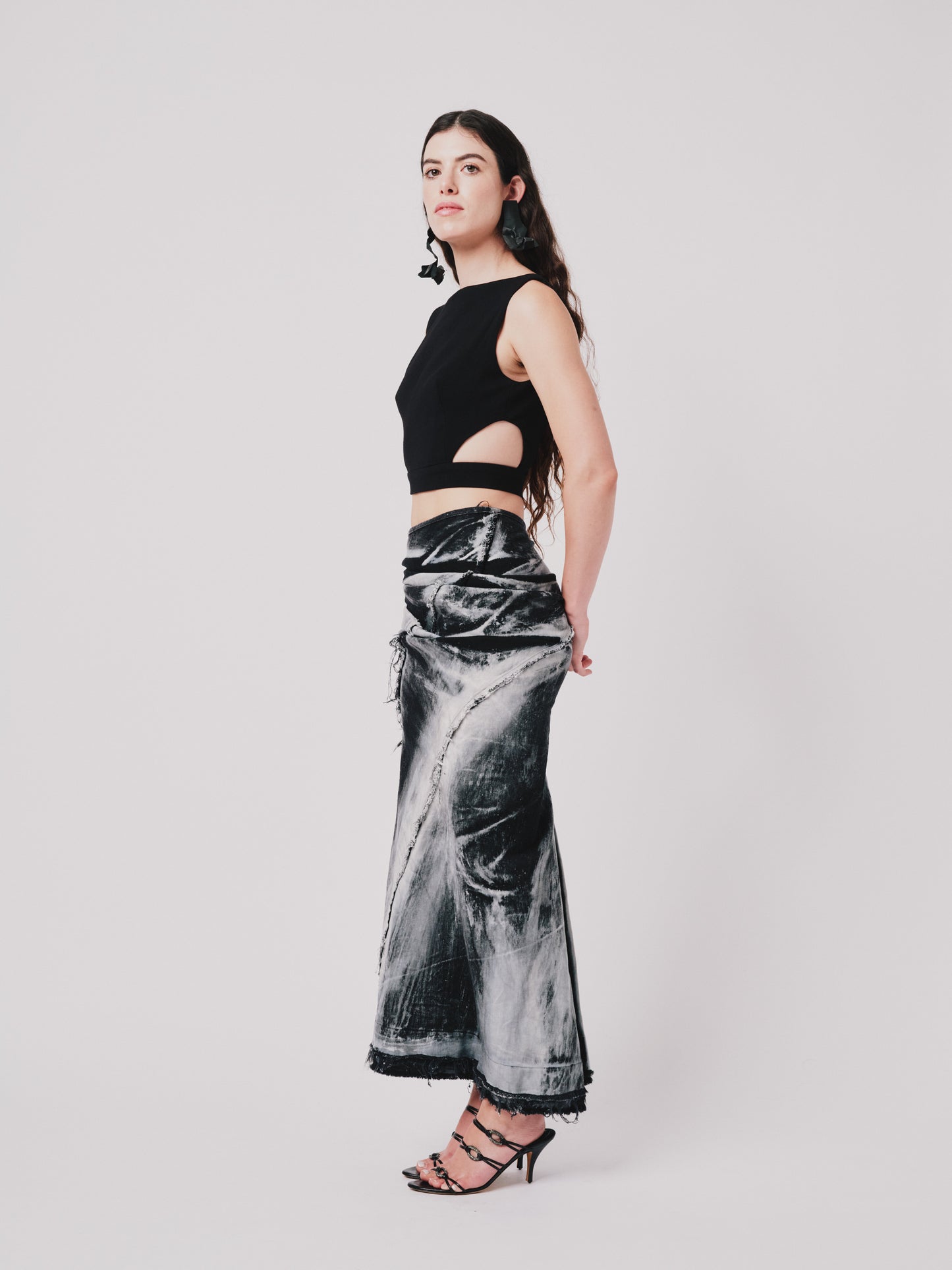 Woman wearing a structured drape skirt in distressed black denim on a plain background. Side view