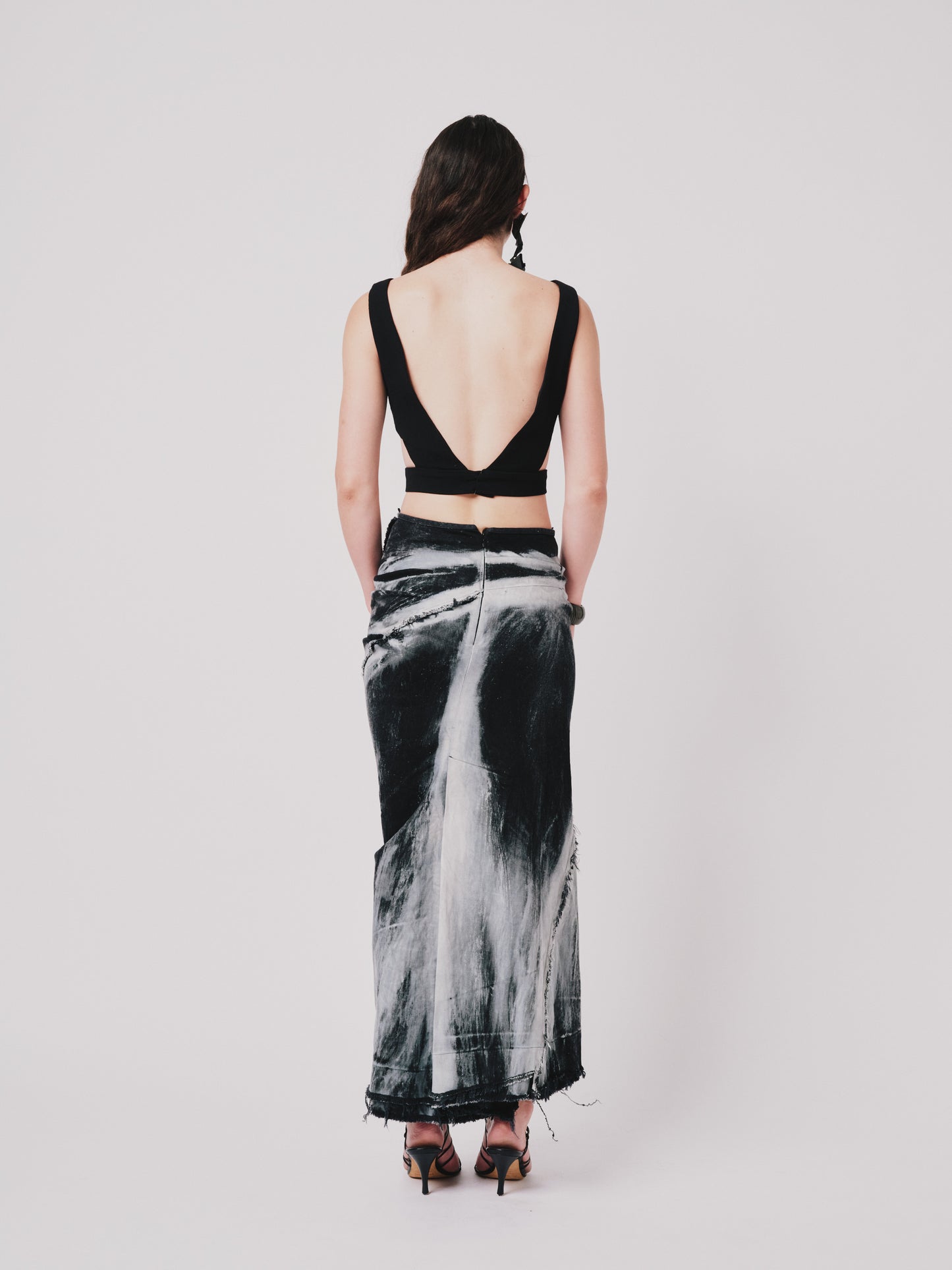 Woman wearing a structured drape skirt in distressed black denim on a plain background. Back view
