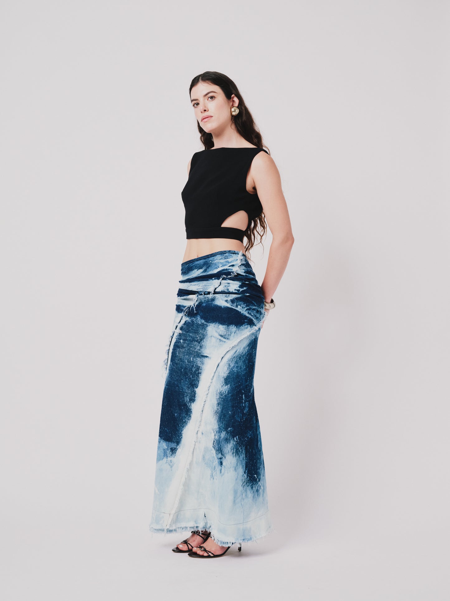 Woman wearing a black top and structured drape skirt in blue denim on a white background. Side view