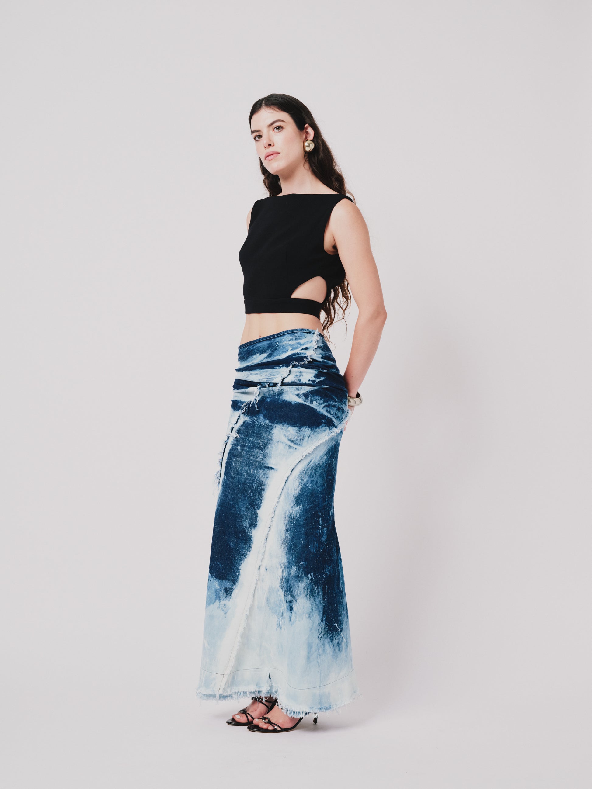 Woman wearing a black top and structured drape skirt in blue denim on a white background. Side view