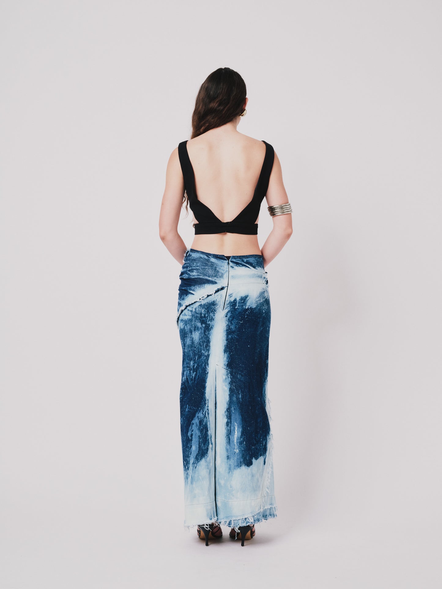 Woman wearing a black top and structured drape skirt in blue denim on a white background. Back view
